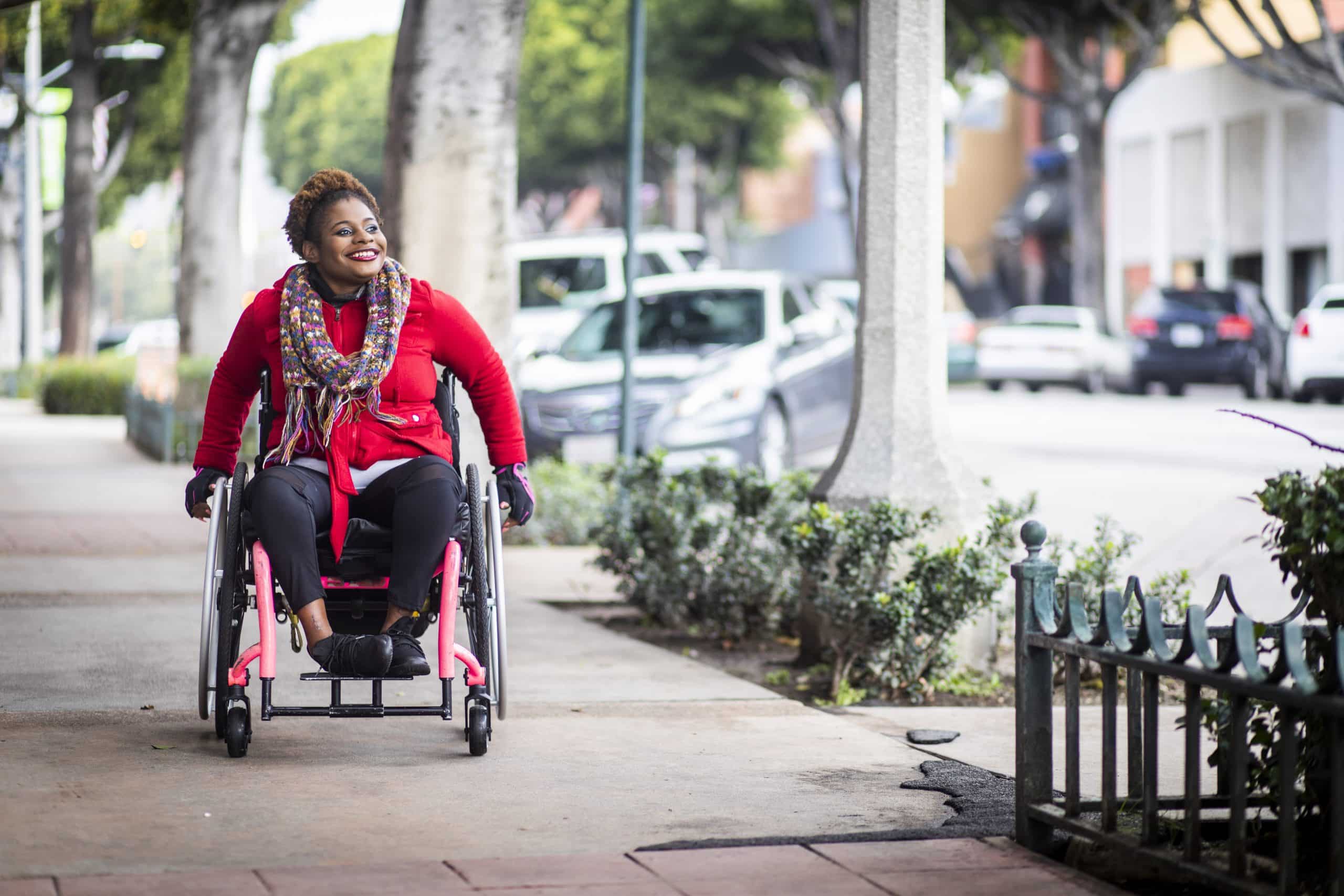 A young black disabled woman with a wheelchair and a bright colored sweater smiling on the sidewalk of a city.