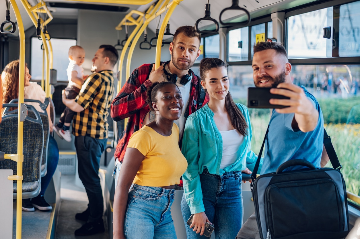 Multiracial smiling group of friends standing in a bus and using smartphone to take a selfie while riding together. Diverse passengers enjoying city ride and taking picture. Public transport.